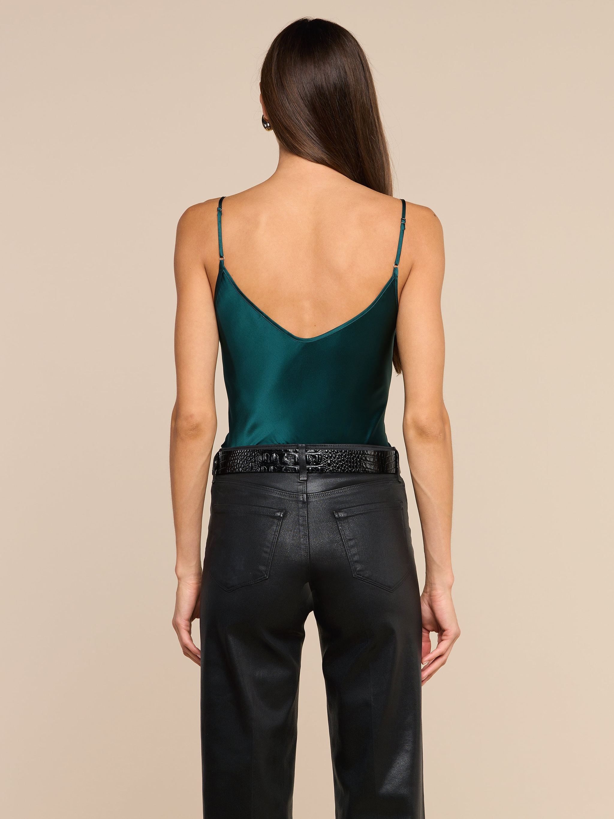 A woman stands with her back to the camera, wearing the Lexi Silk Camisole in dark green—with adjustable straps—and black high-waisted pants. Her straight brown hair cascades down her back, set against a plain light beige background.