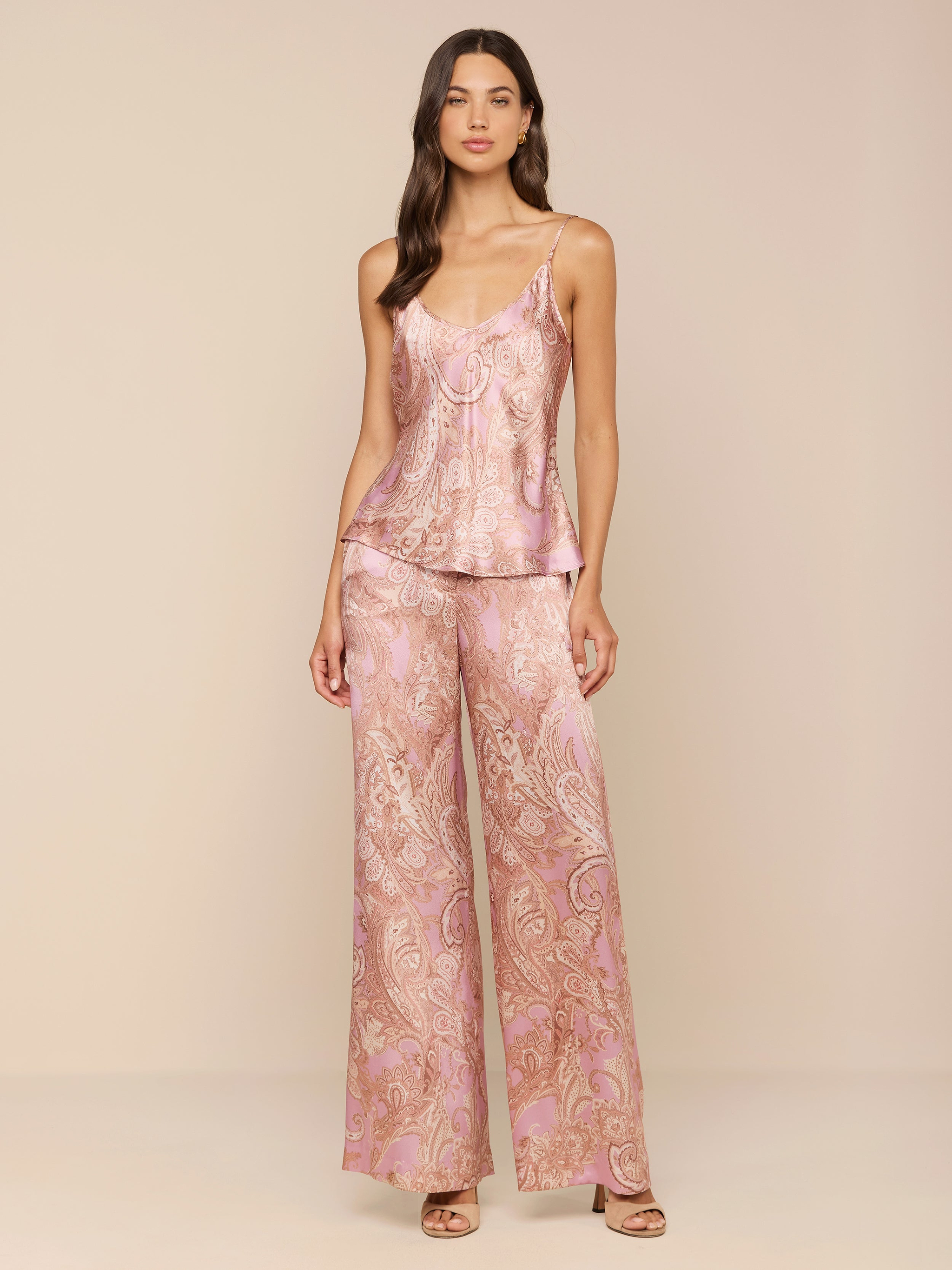 A woman stands against a plain white background wearing the Lexi Silk Camisole in pink paisley satin, featuring adjustable straps, paired with matching wide-leg pajama pants and heeled sandals.