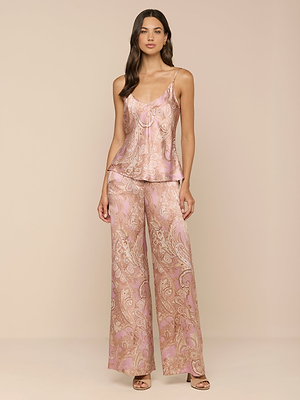 Una donna posa su uno sfondo beige con il Lexi Silk Camisole, un top rosa senza maniche a motivi paisley con spalline regolabili, abbinato a pantaloni a gamba larga e tacchi aperti. I capelli scuri sono sciolti e la donna sembra rilassata.