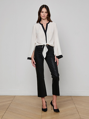 A woman stands indoors in the Charlize Silk Blouse, an ivory tie-front design with black trim, paired with black cropped pants and heels, posing against a plain white wall on a wooden floor.