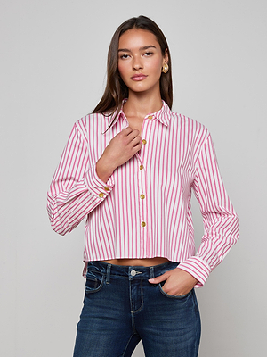 A woman with long brown hair and a neutral expression wears the L'AGENCE Cosette Shirt, a classic tailored organic cotton piece with pink and white stripes, paired with blue jeans, standing against a light gray background.
