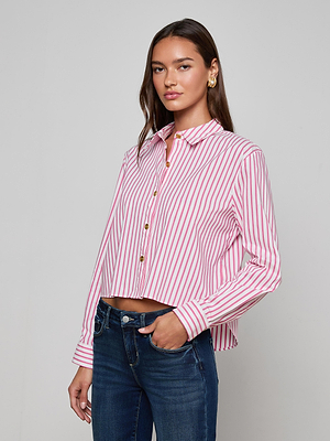A woman with long brown hair models the L'AGENCE Cosette Shirt—a classic tailored pink and white stripe button-up—paired with blue jeans, standing against a plain light gray background.