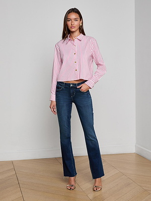 A woman poses indoors in the L'AGENCE Cosette Shirt—an organic cotton, pink and white striped top—paired with blue bootcut jeans and heeled sandals, standing against a plain background with one hand in her pocket.
