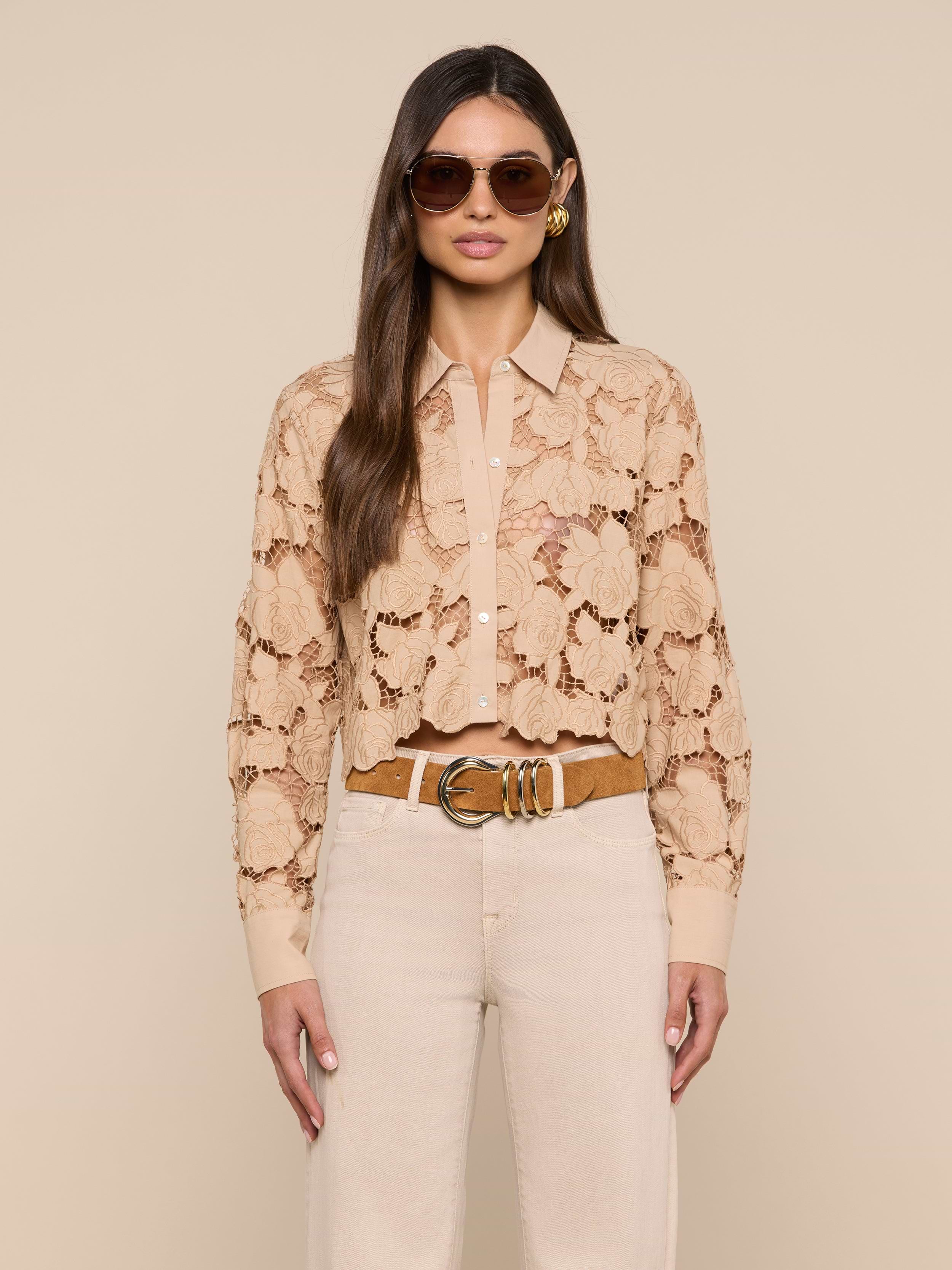 A woman wearing sunglasses and the Seychelle Cropped Eyelet Blouse stands facing forward against a plain light background, paired with cream pants and a brown belt.