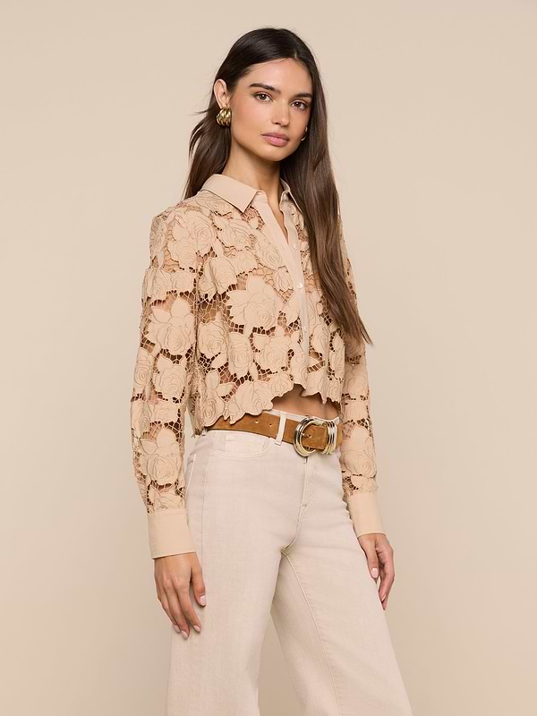 A woman wearing the Seychelle Cropped Eyelet Blouse—a beige floral eyelet top with a cropped silhouette—layered over a collared shirt and paired with high-waisted cream pants and a tan belt, stands against an off-white background.