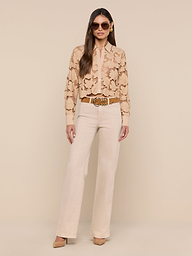 A woman stands against a white background wearing sunglasses, the Seychelle Cropped Eyelet Blouse, high-waisted light beige pants, and tan heels, with her hands in her pockets.