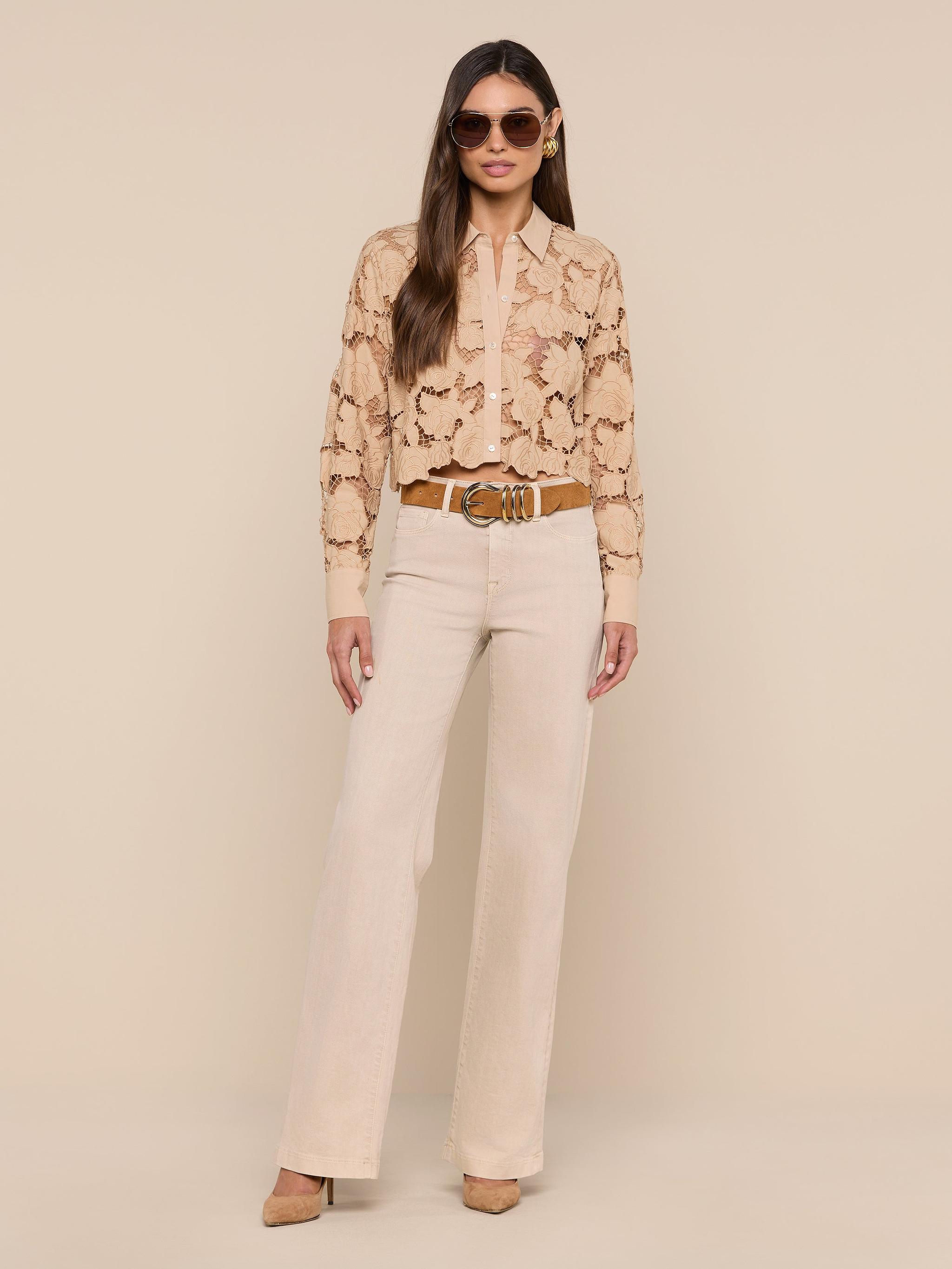 A woman stands against a white background wearing sunglasses, the Seychelle Cropped Eyelet Blouse, high-waisted light beige pants, and tan heels, with her hands in her pockets.