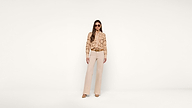 A woman wearing sunglasses, a beige snakeskin-patterned blouse, beige pants, and brown shoes stands against a plain white background.