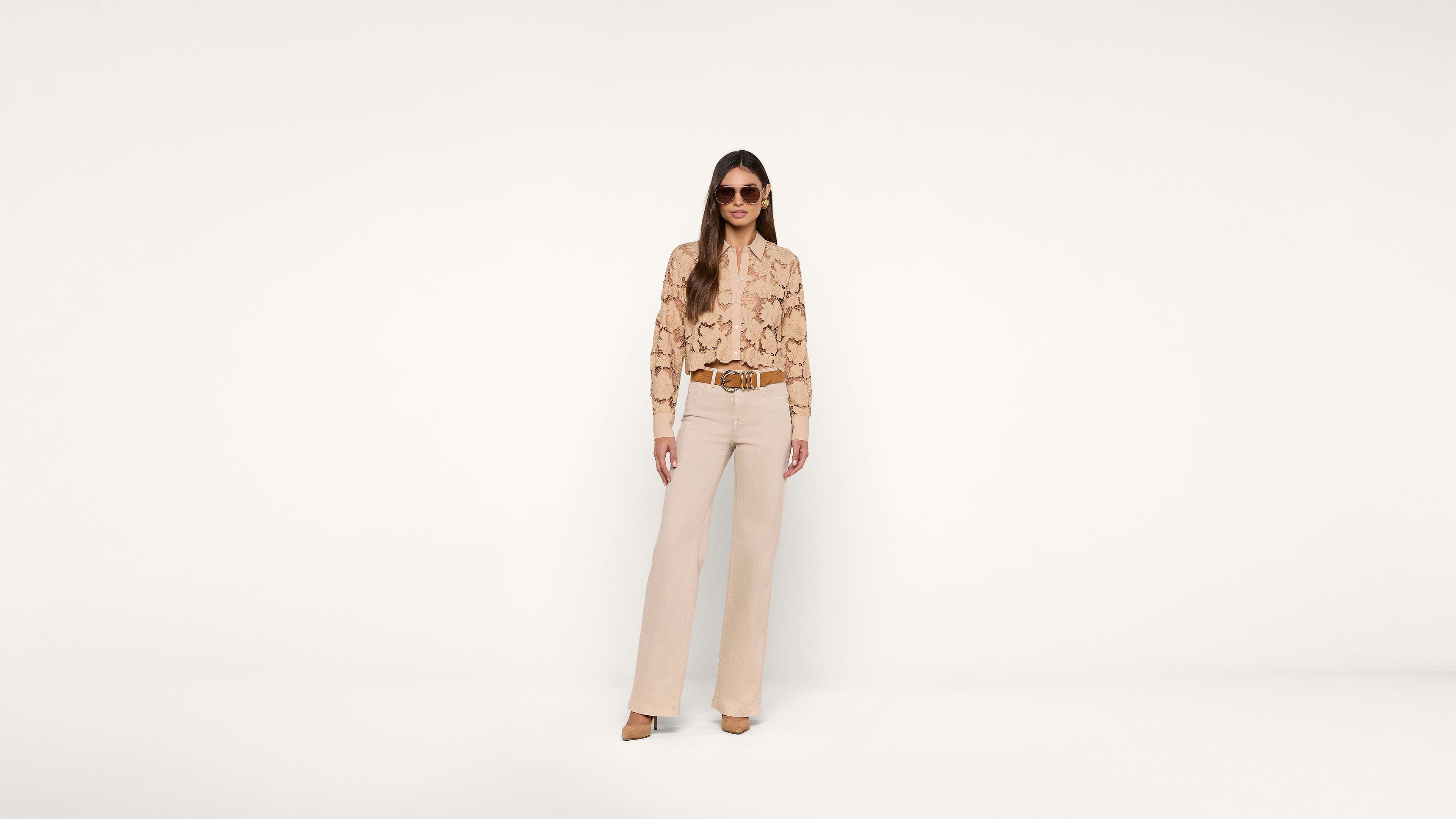 A woman wearing sunglasses, a beige snakeskin-patterned blouse, beige pants, and brown shoes stands against a plain white background.