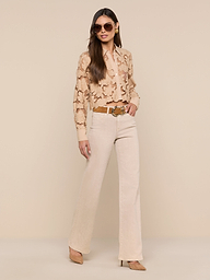 A woman stands against a plain white background, facing forward with one hand in her pocket, wearing beige trousers, a brown belt, heels, sunglasses, and the Seychelle Cropped Eyelet Blouse.