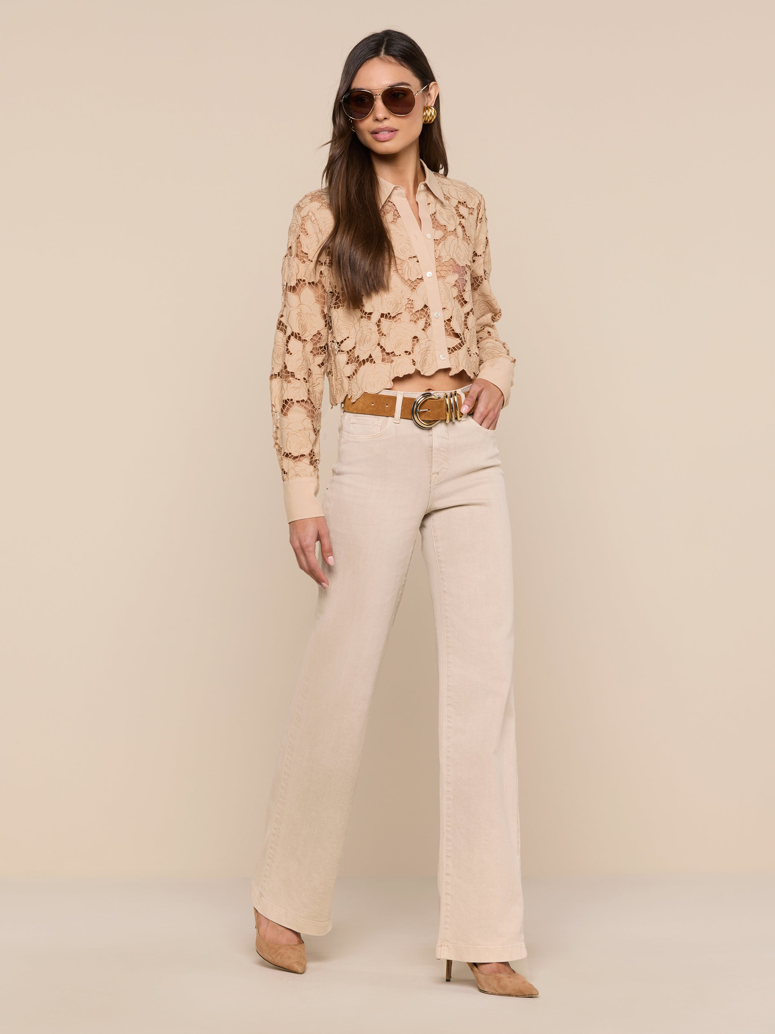 A woman stands against a plain white background, facing forward with one hand in her pocket, wearing beige trousers, a brown belt, heels, sunglasses, and the Seychelle Cropped Eyelet Blouse.