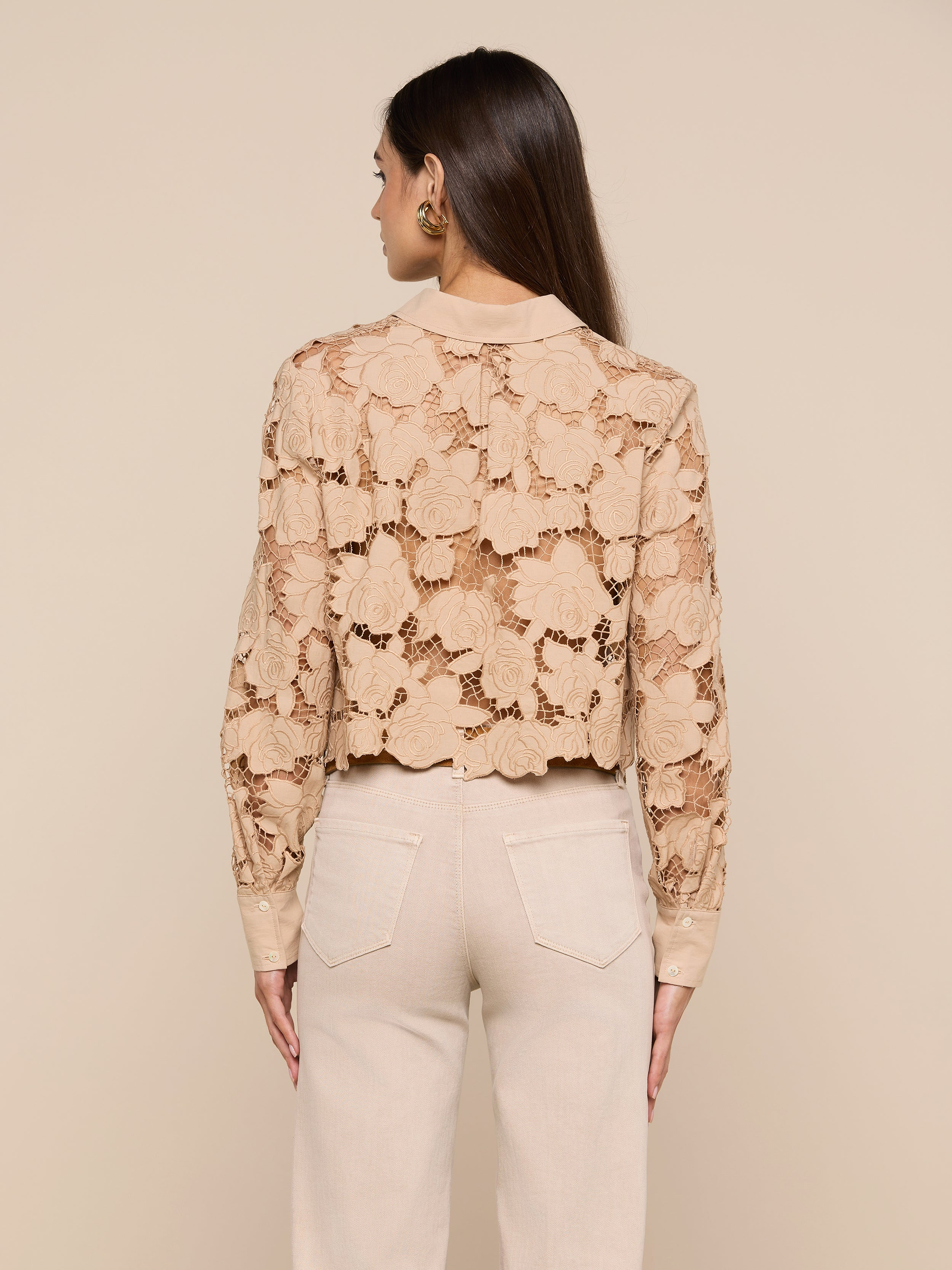 A person with long brown hair is shown from behind, wearing the Seychelle Cropped Eyelet Blouse—beige with floral patterns—and high-waisted beige pants against a plain light background.