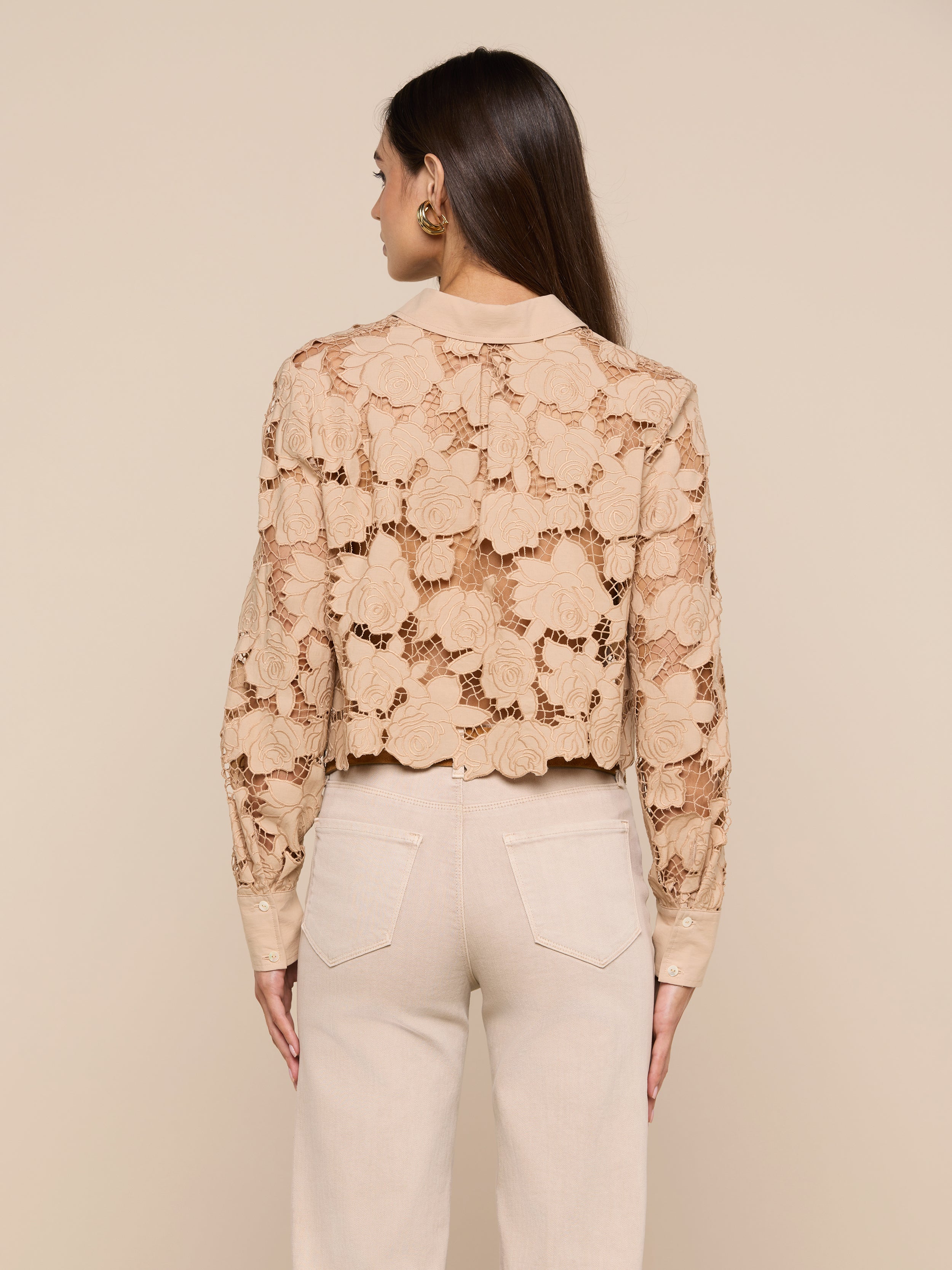 A person with long brown hair is shown from behind, wearing the Seychelle Cropped Eyelet Blouse—beige with floral patterns—and high-waisted beige pants against a plain light background.