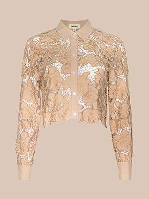 The Seychelle Cropped Eyelet Blouse is a beige long-sleeve button-up top with an intricate floral lace pattern, pointed collar, and semi-sheer design that subtly reveals a white background for an elegant, delicate look.