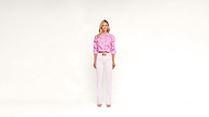 A woman stands against a plain white background, wearing a pink floral blouse, white pants, and white shoes. She has blonde hair and a neutral expression.