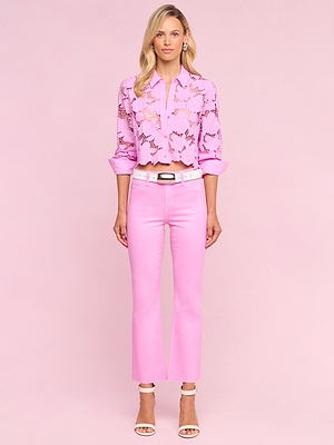 A woman with loose waves stands against a light pink background, wearing the Seychelle Cropped Eyelet Blouse from the Legally Blonde capsule collection, paired with pink pants, a white belt, and white open-toe heels.