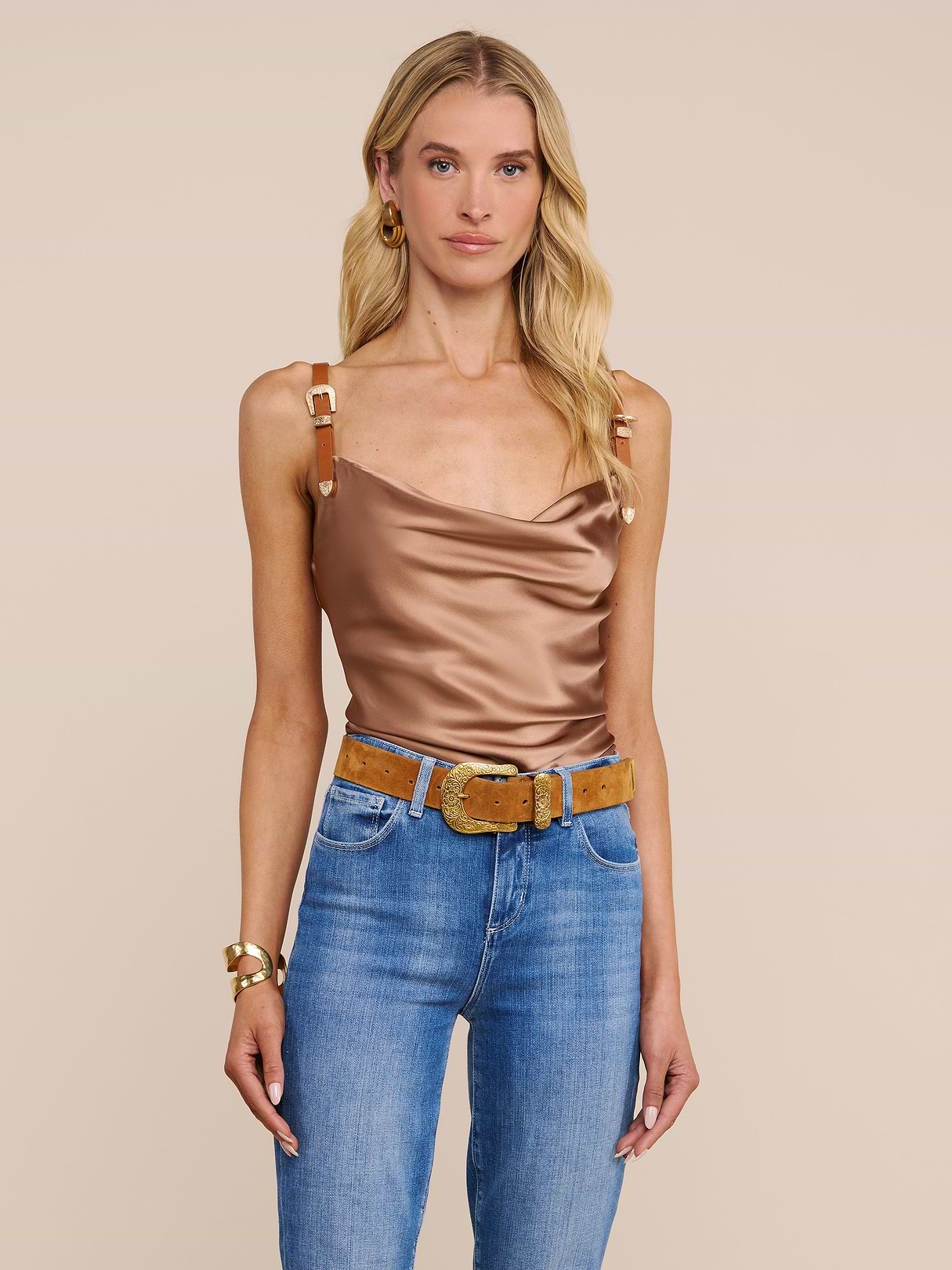 Una donna con lunghi capelli biondi indossa la canotta Lux Silk Buckle-Strap, abbinandola a jeans blu, una cintura marrone con fibbia decorativa, grandi orecchini a cerchio e bracciali dorati su uno sfondo beige.
