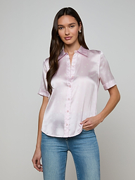 A woman wearing the Ellah Silk Blouse in light pink, featuring short sleeves and a collar, paired with blue jeans, stands against a plain light background.