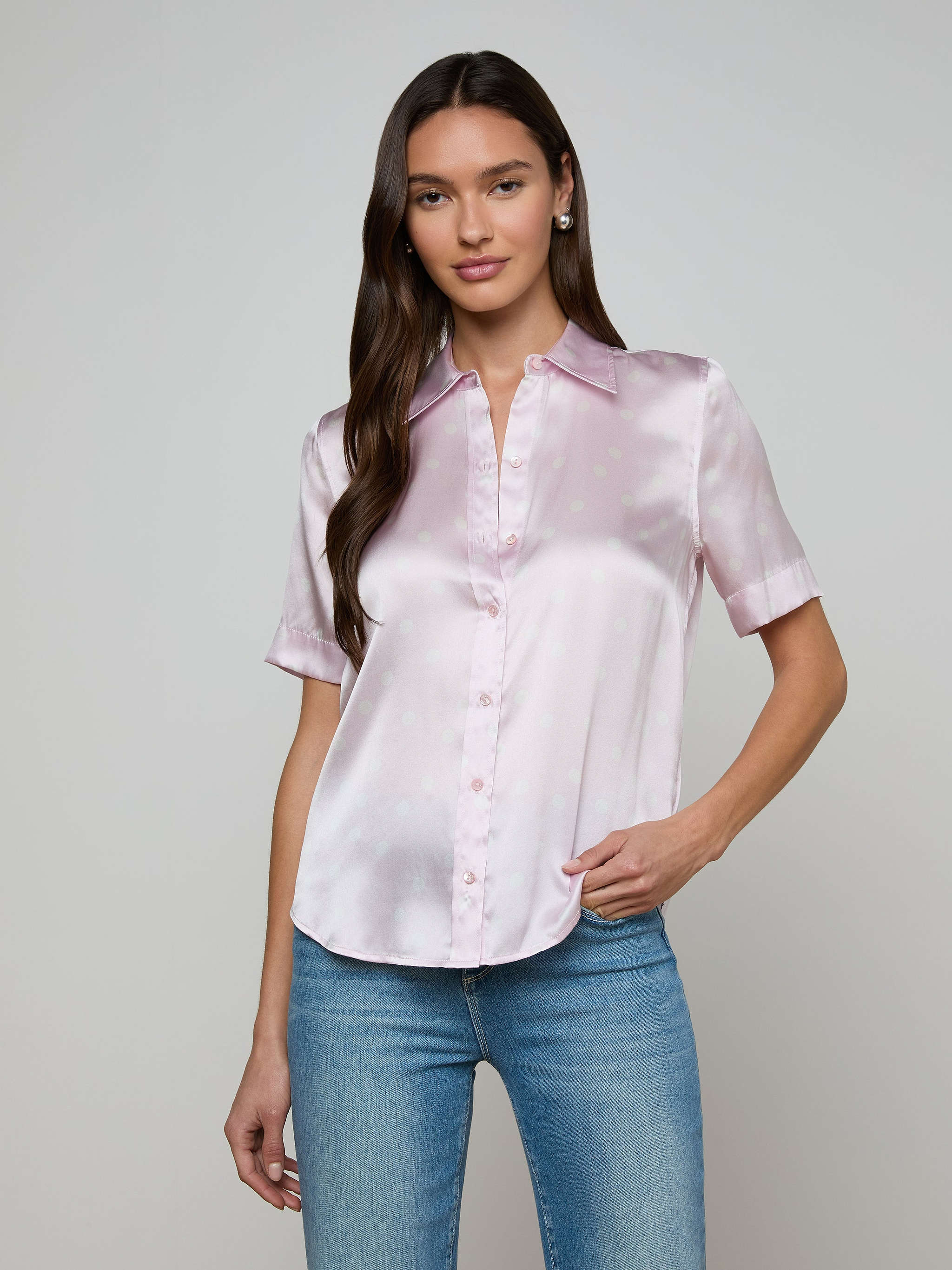 A woman wearing the Ellah Silk Blouse in light pink, featuring short sleeves and a collar, paired with blue jeans, stands against a plain light background.