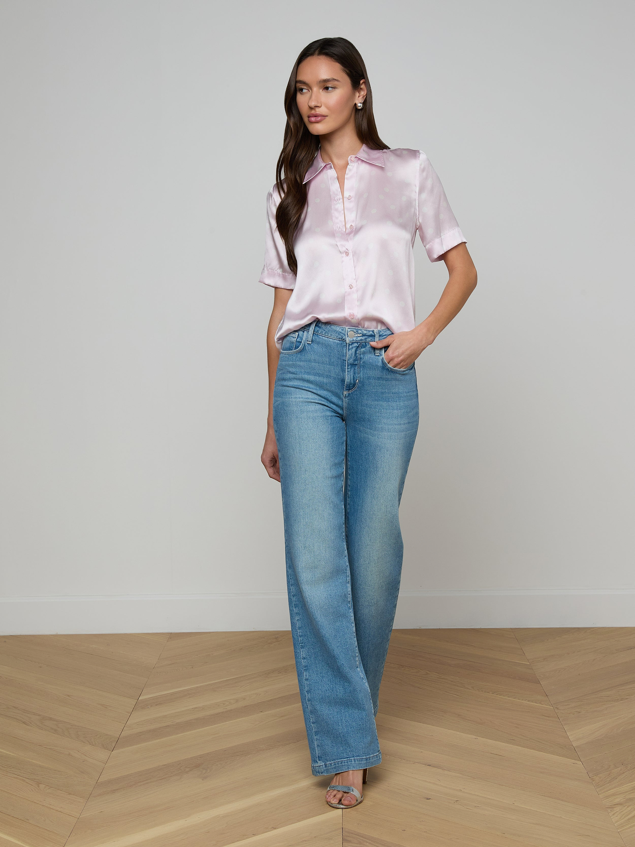 A woman stands against a plain white background wearing the Ellah Silk Blouse in light pink, paired with blue high-waisted wide-leg jeans and open-toe shoes.