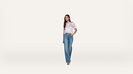 A woman wearing a light pink short-sleeve blouse and blue jeans stands against a plain white background, with one hand in her pocket and a neutral expression.