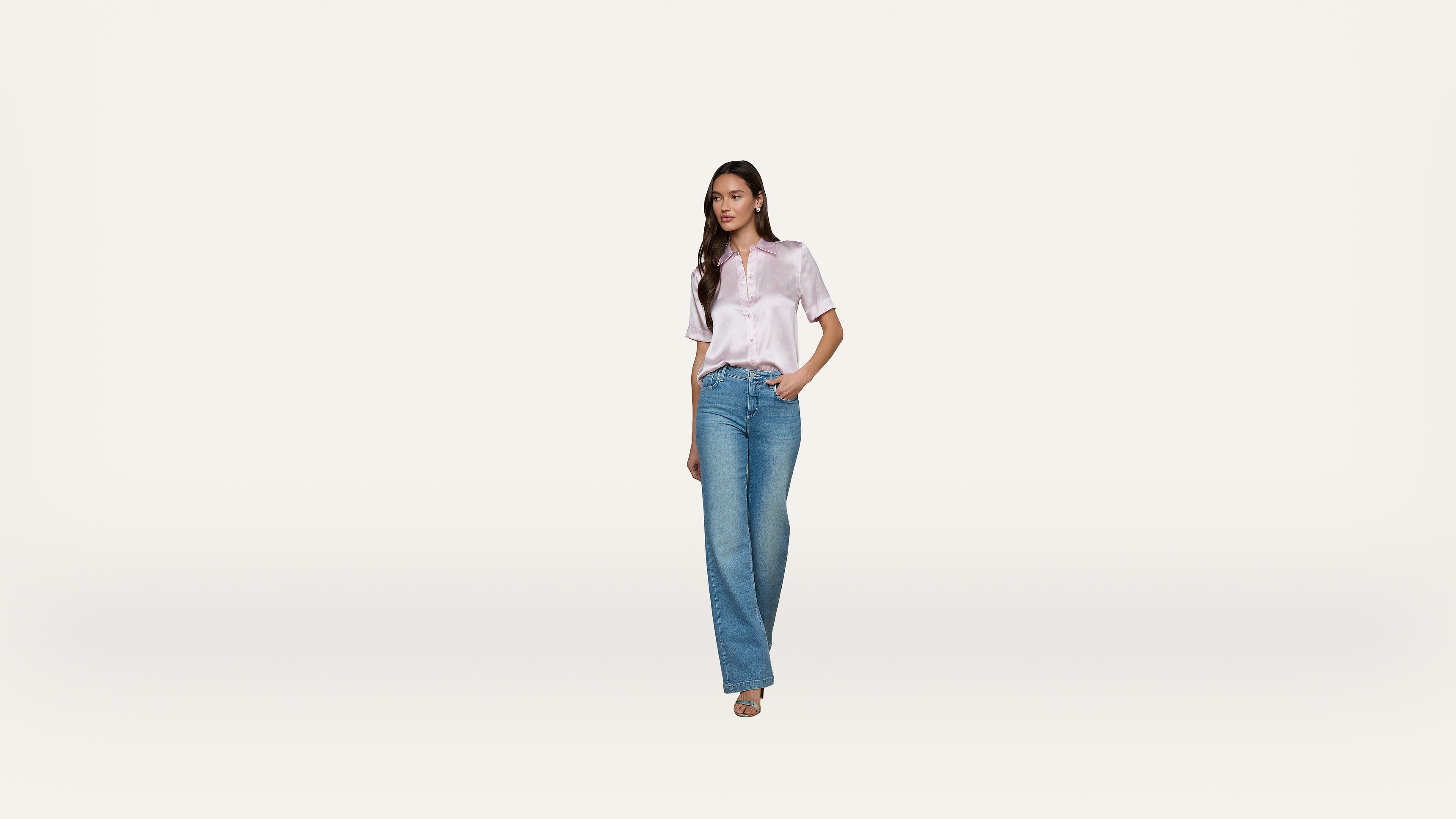 A woman wearing a light pink short-sleeve blouse and blue jeans stands against a plain white background, with one hand in her pocket and a neutral expression.