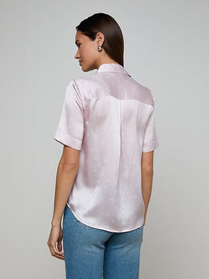 A woman with straight brown hair wears the Ellah Silk Blouse, a light pink, short-sleeve top with a subtle polka dot pattern, paired with light blue jeans. She stands with her back to the camera against a plain light background.
