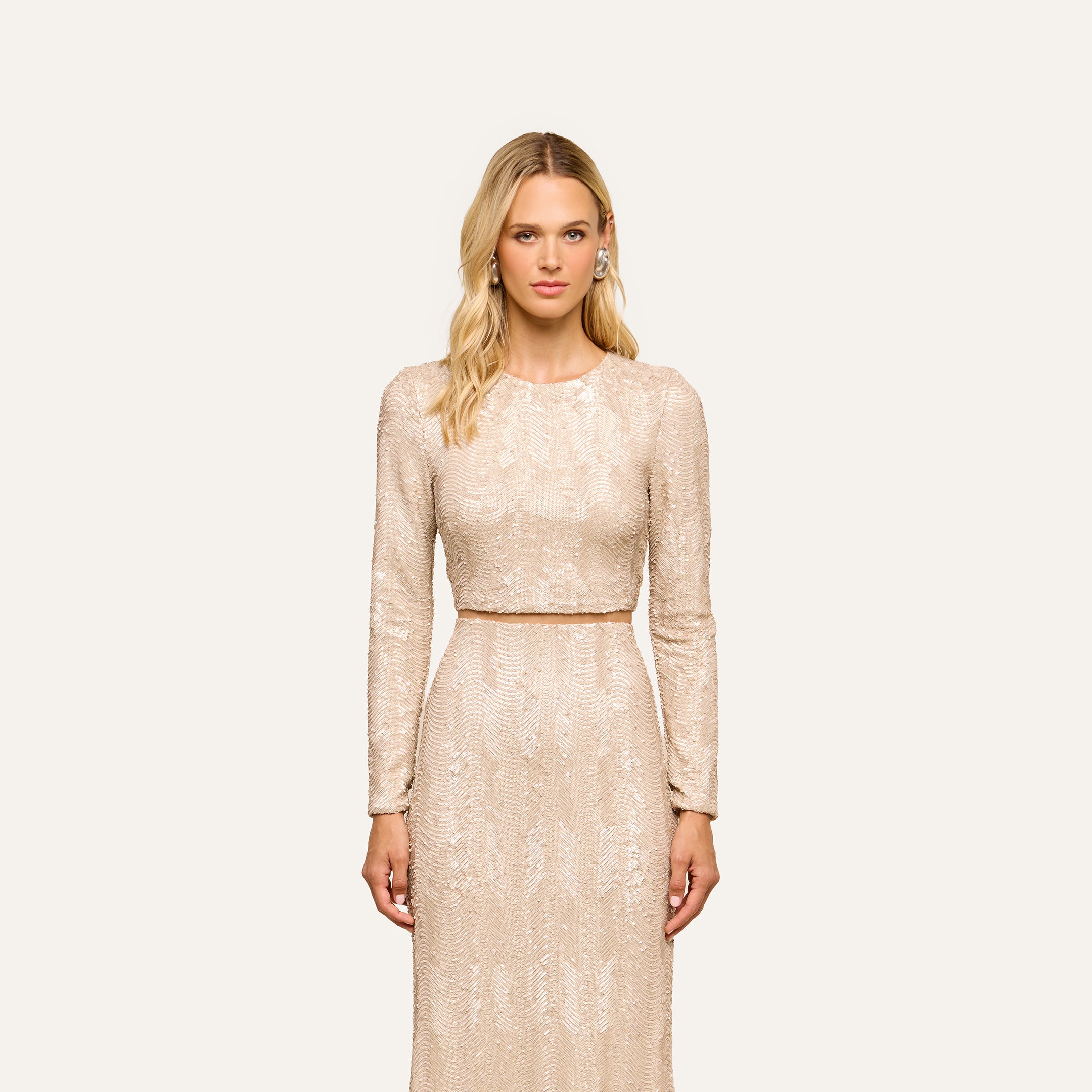 A woman with blonde hair wears the Rogan Sequin Top, a long-sleeved piece embellished with champagne sequins for a sophisticated, party-ready look. She stands facing forward against a plain, light background.