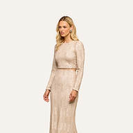 A woman with long blonde hair stands against a plain off-white background, wearing the party-ready Rogan Sequin Top—a light beige, long-sleeve crop top—paired with a matching floor-length skirt.