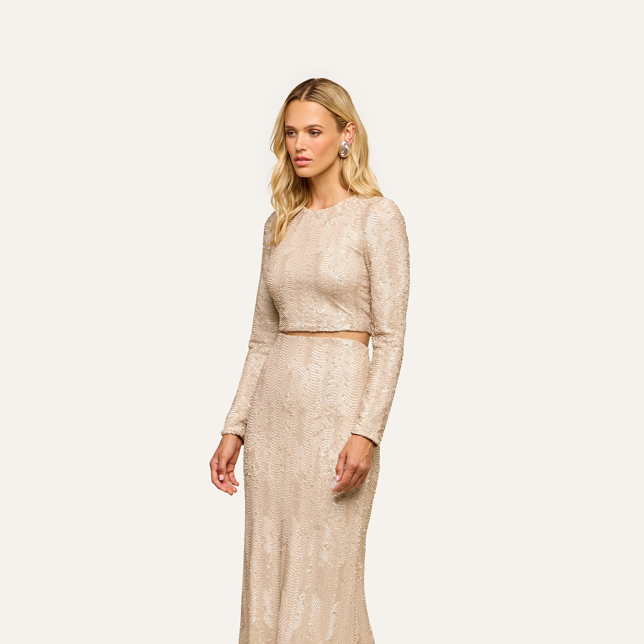 A woman with long blonde hair stands against a plain off-white background, wearing the party-ready Rogan Sequin Top—a light beige, long-sleeve crop top—paired with a matching floor-length skirt.