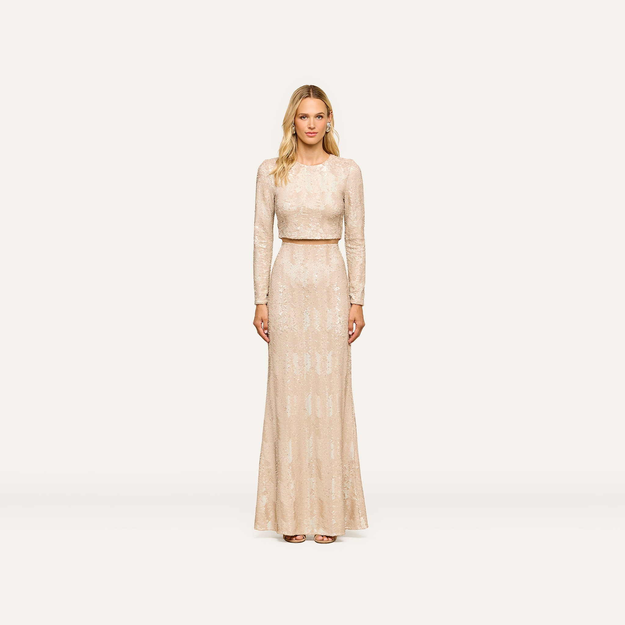 A woman wearing the Rogan Sequin Top, a light beige long-sleeve blouse embellished with champagne sequins, stands facing forward against a plain, light background for a party-ready look.
