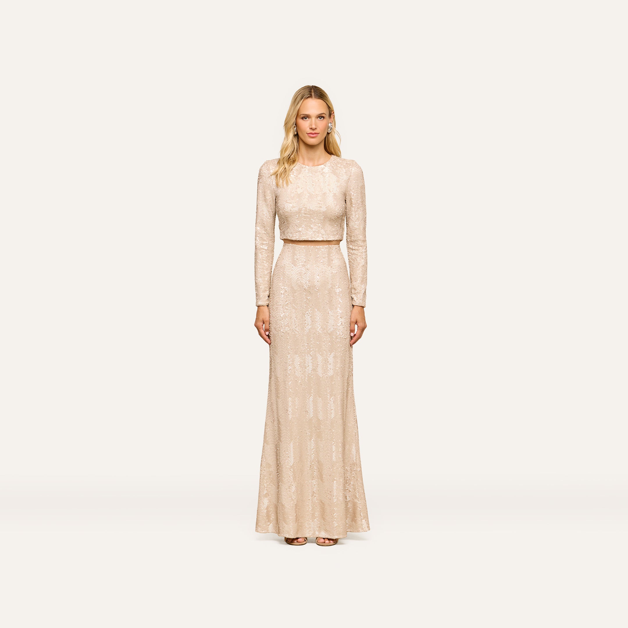 A woman wearing the Rogan Sequin Top, a light beige long-sleeve blouse embellished with champagne sequins, stands facing forward against a plain, light background for a party-ready look.