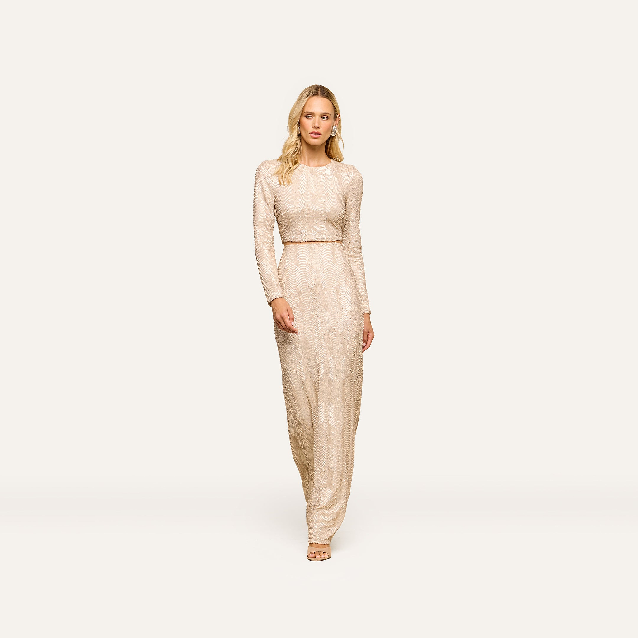 Wearing the Rogan Sequin Top, a woman with long blonde hair pairs it with nude heels and stands against a plain, light background.