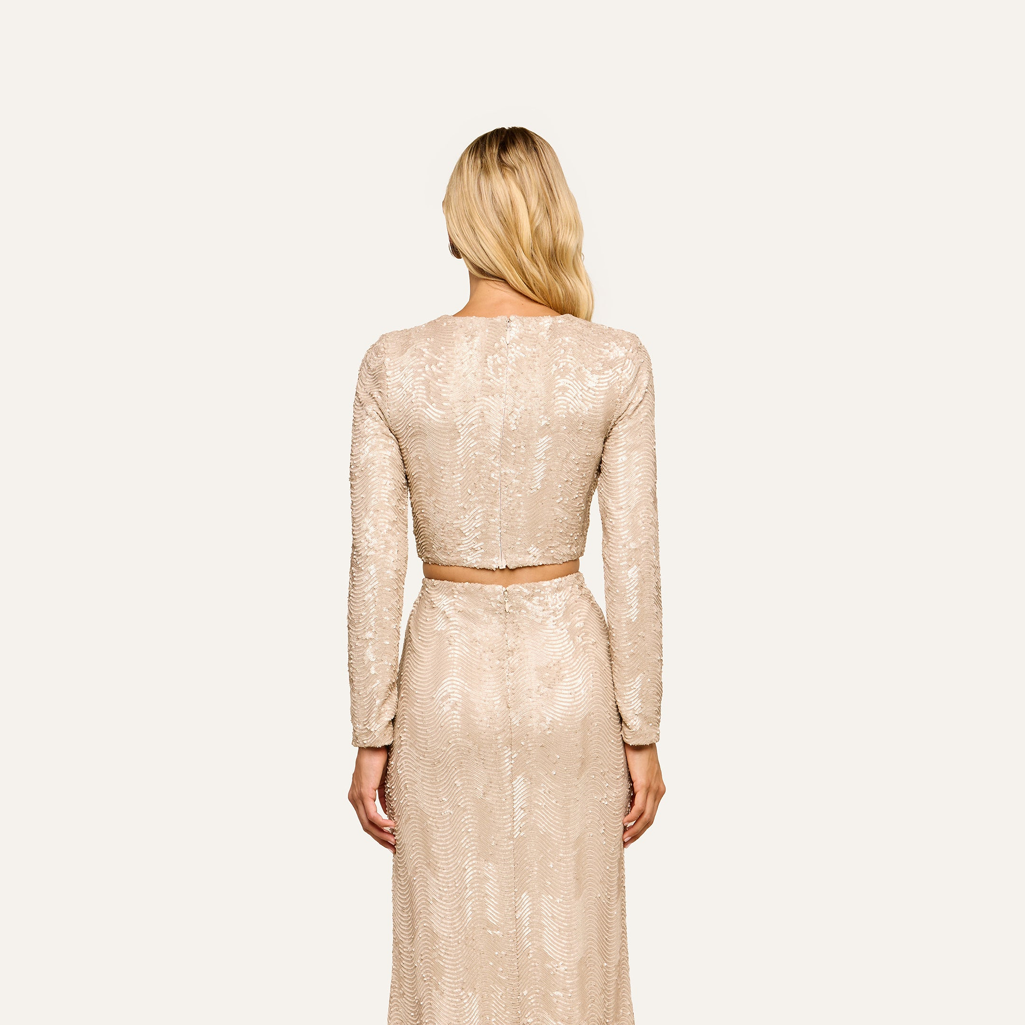 A woman with long blonde hair, seen from behind, wears the Rogan Sequin Top—a party-ready, long-sleeved champagne sequin crop top—paired with a high-waisted skirt against a plain, light background.