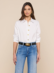Une femme aux longs cheveux bruns porte le modèle Ellery Button-Down Shirt en blanc, décontracté et rentré dans un jean bleu avec une ceinture noire, sur un fond beige uni.