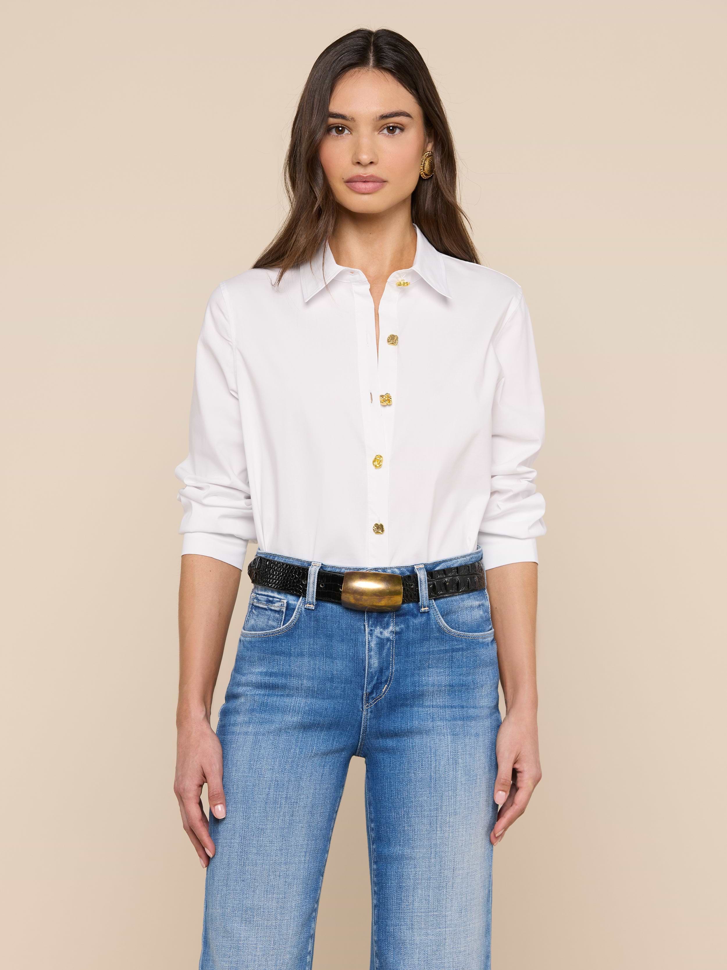 Une femme aux longs cheveux bruns porte le modèle Ellery Button-Down Shirt en blanc, décontracté et rentré dans un jean bleu avec une ceinture noire, sur un fond beige uni.