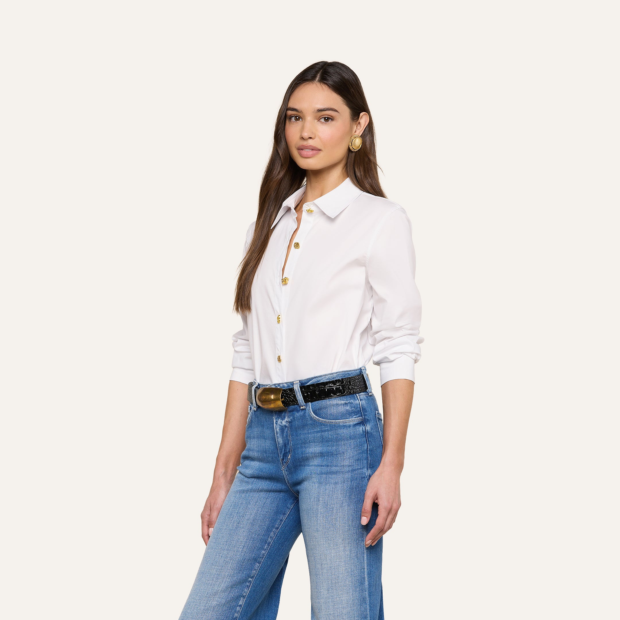 Une femme aux longs cheveux bruns porte la chemise Ellery Button-Down - un mélange de coton biologique à la coupe décontractée - avec un jean bleu et des boucles doreilles en or. Elle se tient debout sur un fond beige, regardant lappareil photo avec un léger sourire.