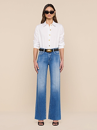 Une femme portant la chemise Ellery Button-Down, un jean bleu à taille haute, une ceinture noire, des sandales noires à talons hauts et des lunettes de soleil se tient debout sur un fond beige uni.