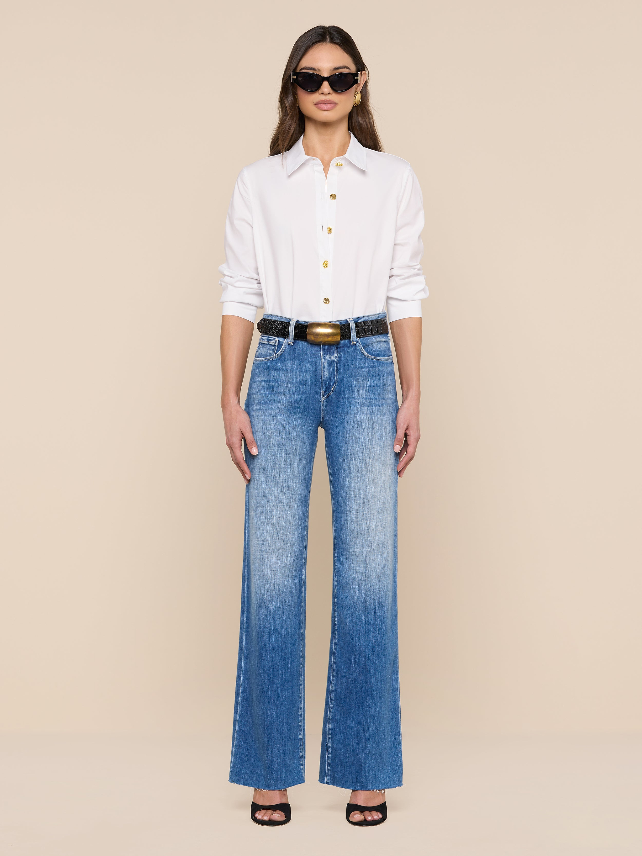 Une femme portant la chemise Ellery Button-Down, un jean bleu à taille haute, une ceinture noire, des sandales noires à talons hauts et des lunettes de soleil se tient debout sur un fond beige uni.