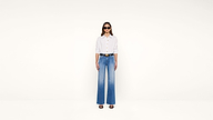 A woman wearing sunglasses, a white button-up shirt, blue high-waisted wide-leg jeans, and black heels stands against a plain white background.