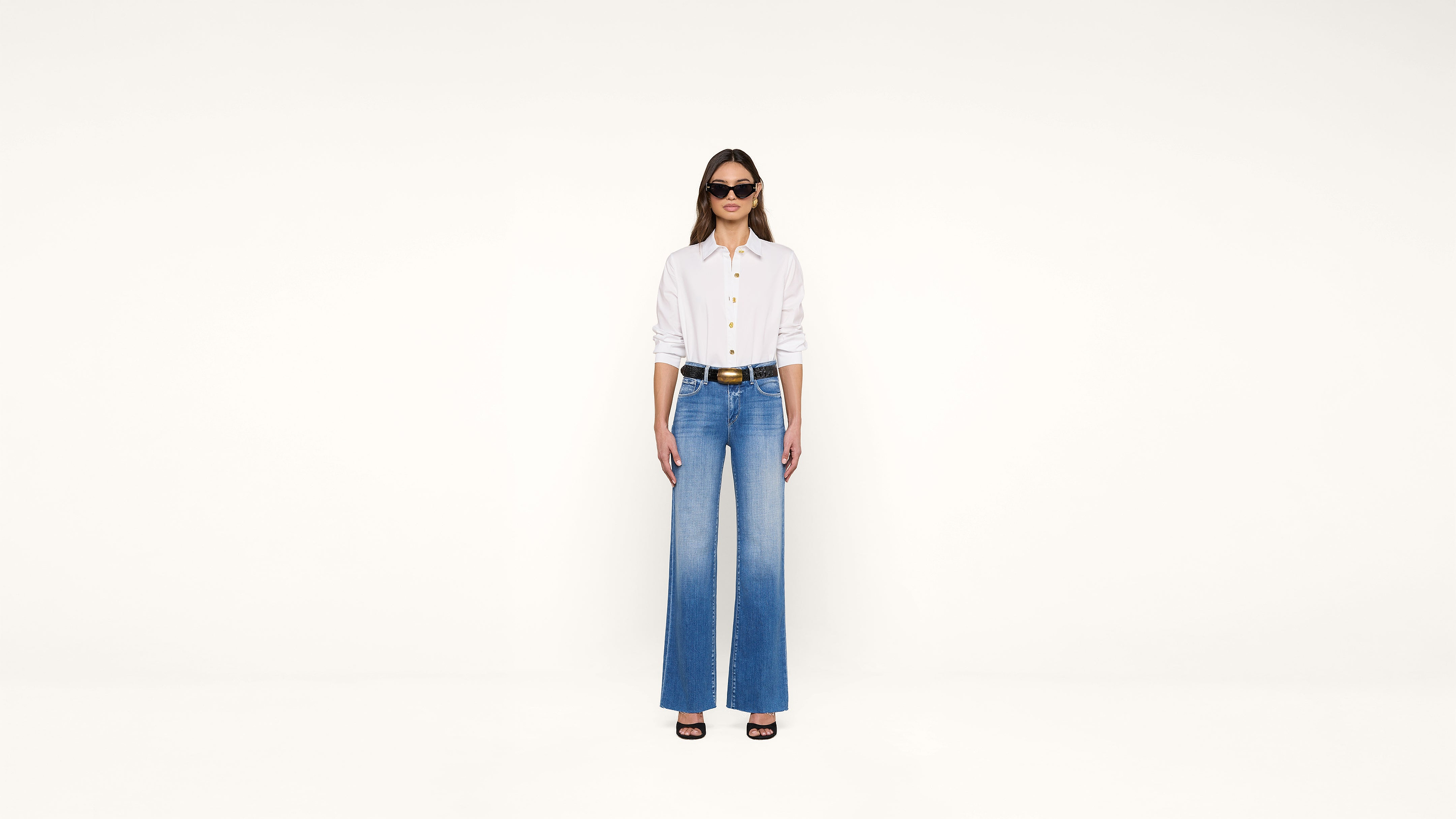 A woman wearing sunglasses, a white button-up shirt, blue high-waisted wide-leg jeans, and black heels stands against a plain white background.