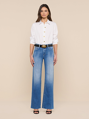 A woman stands against a beige background, facing forward, wearing the Ellery Button-Down Shirt in white, blue high-waisted wide-leg jeans, a black belt with a gold buckle, and black open-toe heels. Her hair is down.