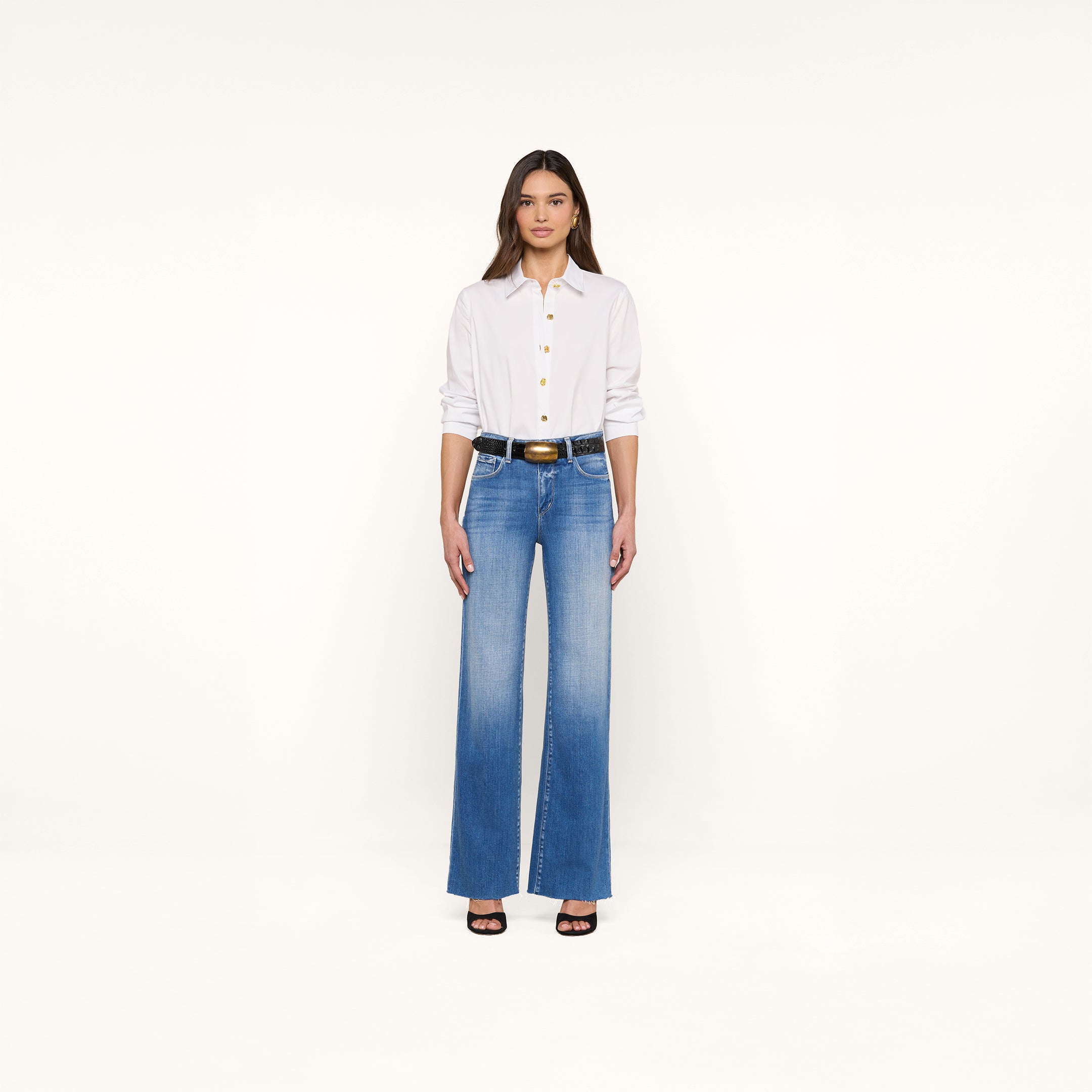 Une femme se tient sur un fond beige, de face, vêtue de la chemise blanche Ellery Button-Down Shirt, dun jean bleu à taille haute, dune ceinture noire à boucle dorée et de talons noirs à bouts ouverts. Ses cheveux sont lâchés.