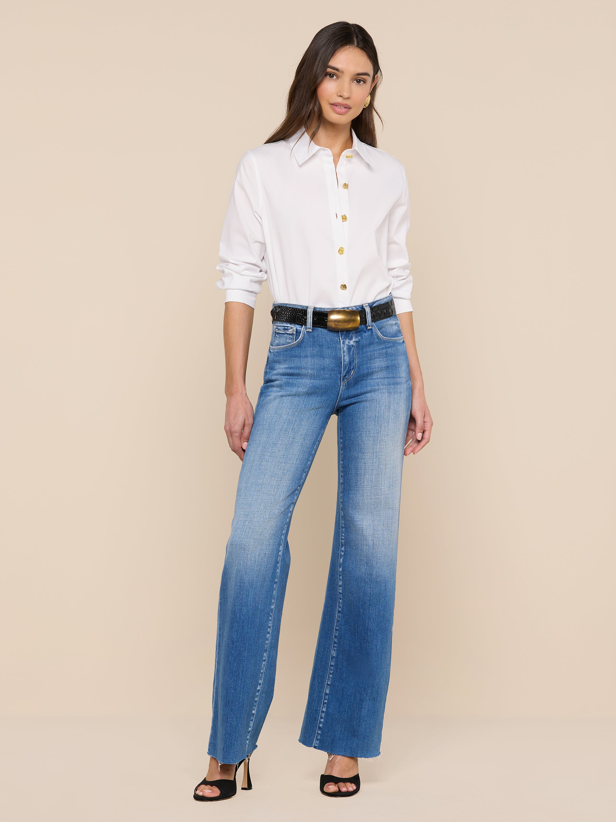 Sur un fond beige, une femme porte la chemise blanche Ellery Button-Down Shirt, associée à un jean bleu à taille haute, à une ceinture noire à boucle dorée et à des sandales noires à talons hauts.