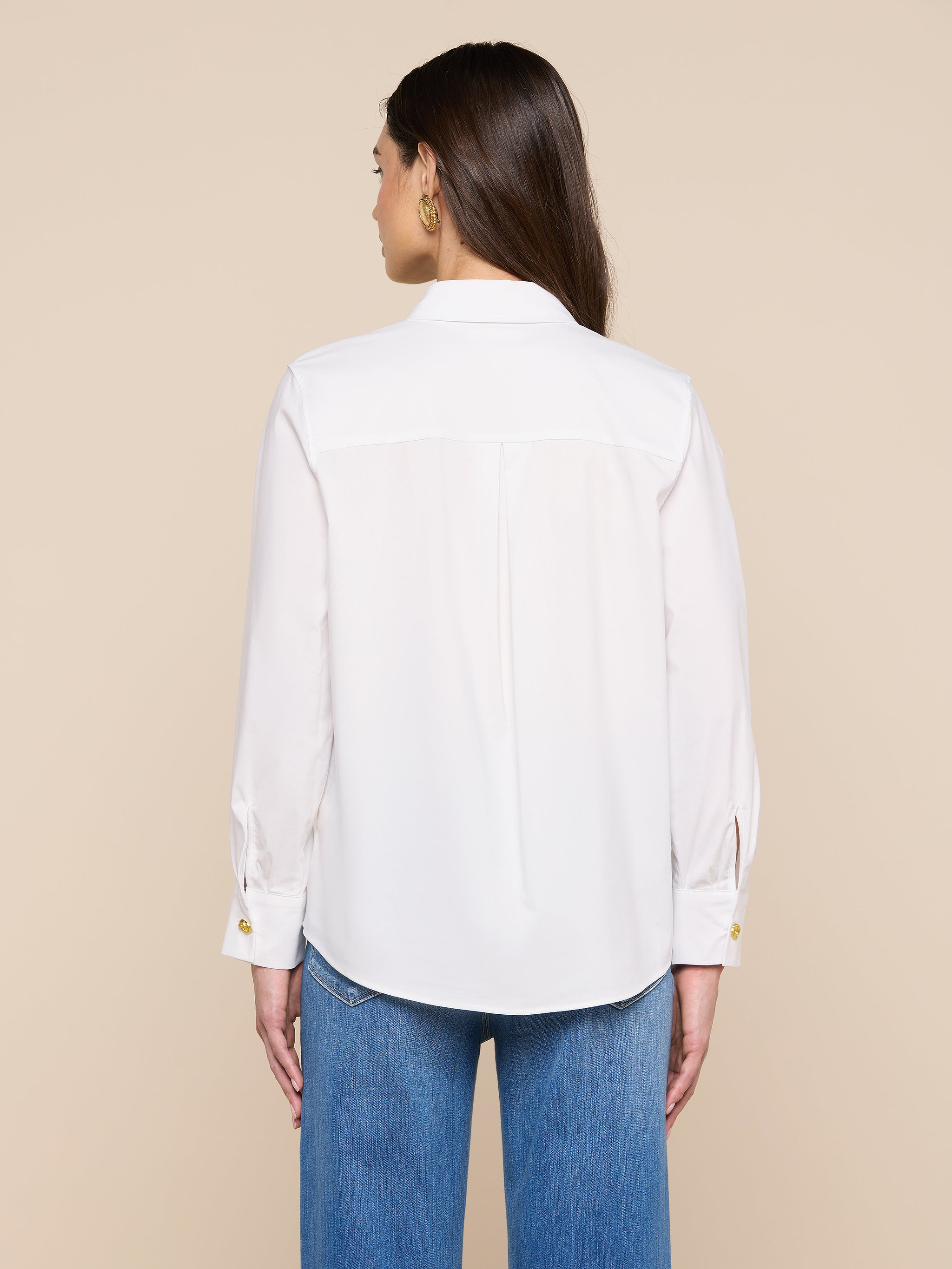 Une femme aux longs cheveux bruns, portant des boucles doreilles en or, un jean bleu et la chemise blanche Ellery Button-Down Shirt se tient de face sur un fond beige.