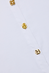 La chemise boutonnée Ellery se compose dun tissu blanc en coton biologique mélangé et dune rangée diagonale de trois boutons dorés uniques, chacun avec un motif abstrait ou en forme de nœud, mettant en valeur des détails texturés et une coupe décontractée.