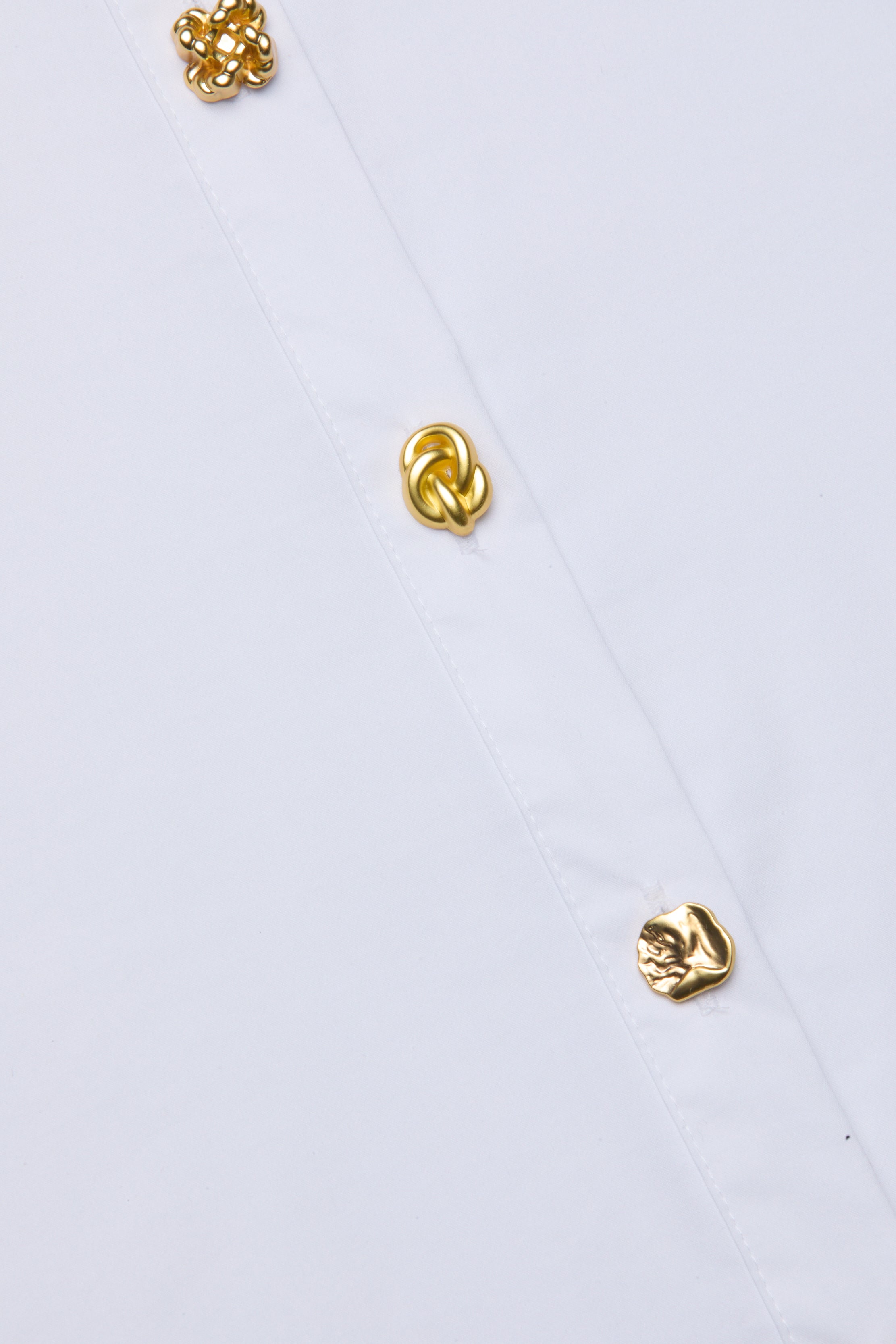 La chemise boutonnée Ellery se compose dun tissu blanc en coton biologique mélangé et dune rangée diagonale de trois boutons dorés uniques, chacun avec un motif abstrait ou en forme de nœud, mettant en valeur des détails texturés et une coupe décontractée.