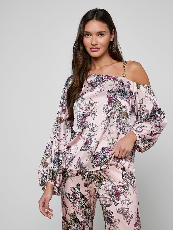 A woman with long brown hair stands against a plain light background, posing with one hand in her pocket while wearing the Asami Chain One-Shoulder Blouse in pink, paired with matching wide-leg floral pants that feature a subtle chain accent.