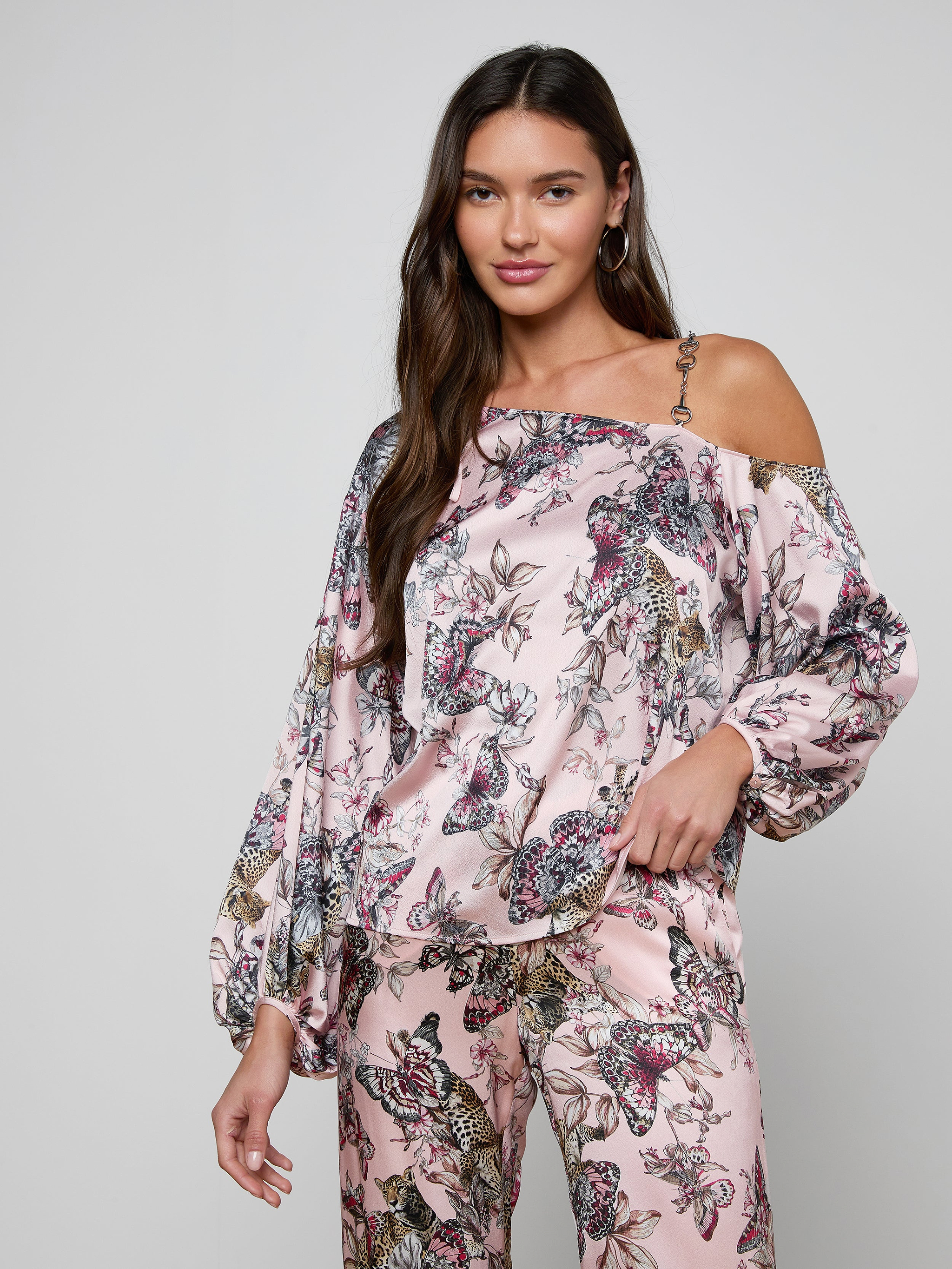A woman with long brown hair stands against a plain light background, posing with one hand in her pocket while wearing the Asami Chain One-Shoulder Blouse in pink, paired with matching wide-leg floral pants that feature a subtle chain accent.