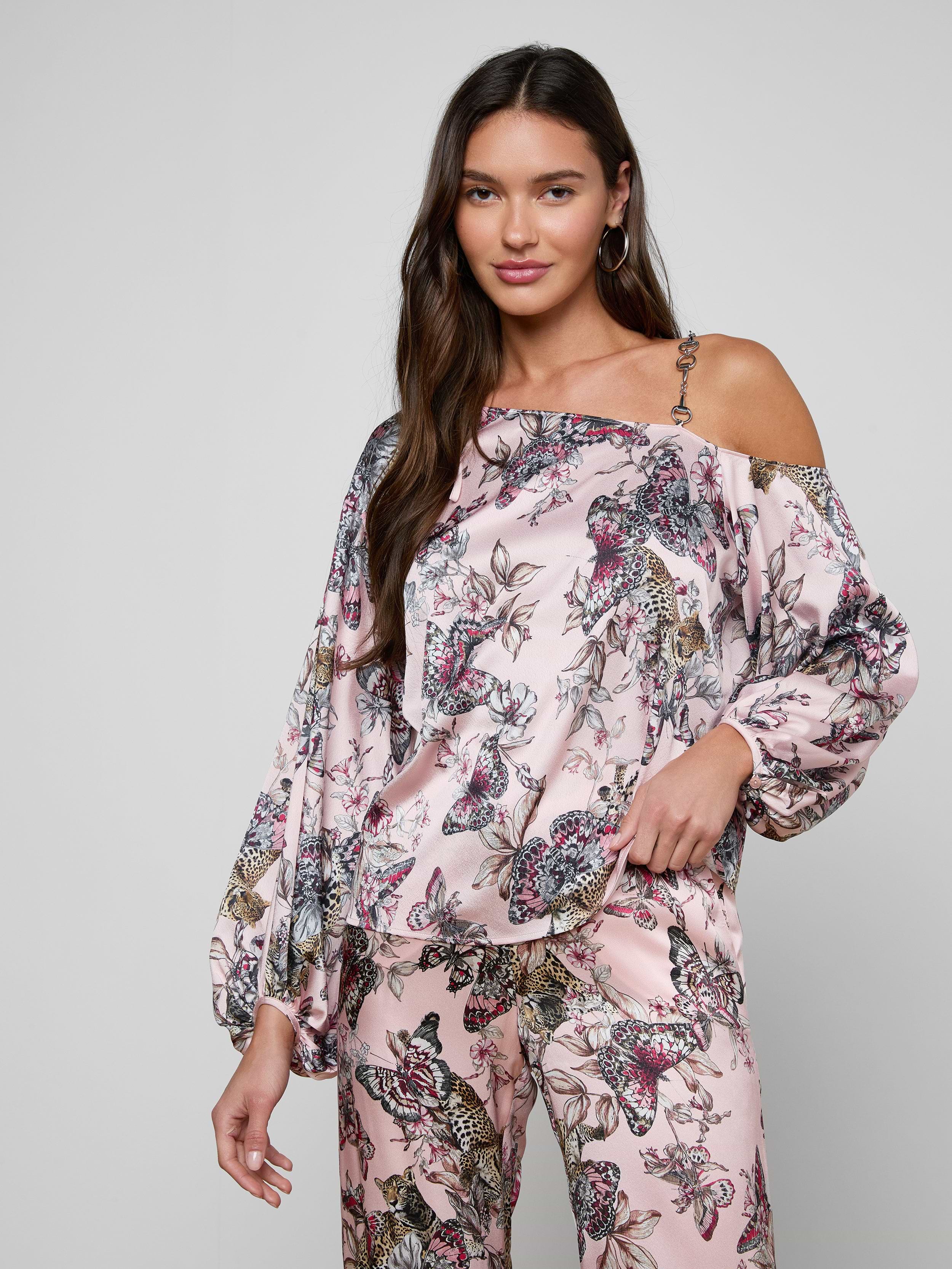 A woman with long brown hair stands against a plain light background, posing with one hand in her pocket while wearing the Asami Chain One-Shoulder Blouse in pink, paired with matching wide-leg floral pants that feature a subtle chain accent.